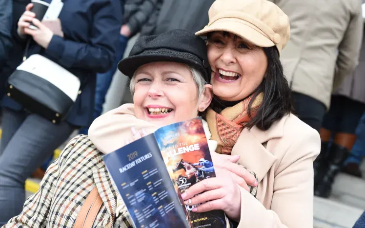 Two racegoers laughing and posing for the camera with their racecard